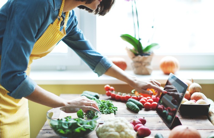 Female in yellow apron scrolling through iPad while handling vegetables in a kitchen