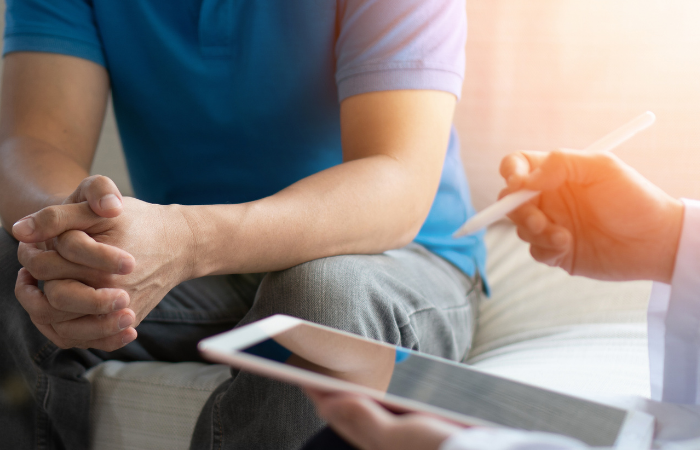 Unidentifiable male patient sitting with hands clasped, waiting for doctor to complete a note on an iPad