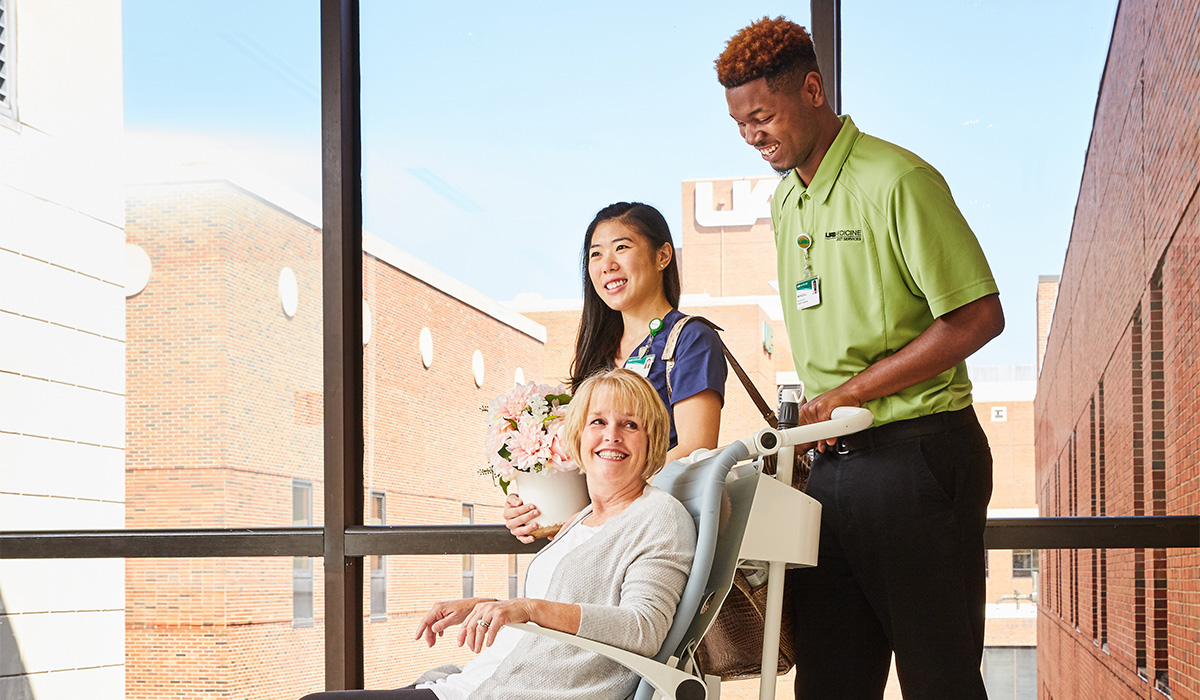 Male African American UAB employee pushing a female caucasian patient in a wheelchair, Asian female employee walking beside them